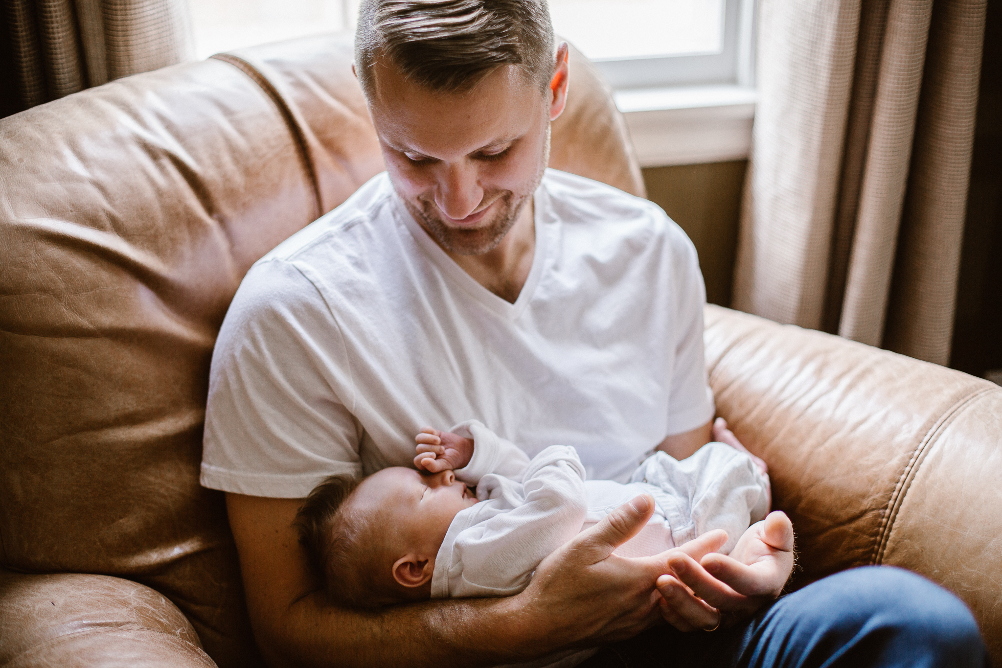 New father cradling his baby boy in a leather chair by the window.